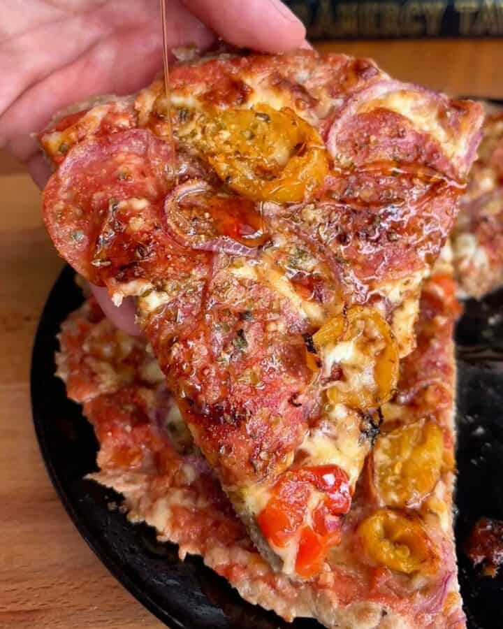 a slice of crispy hot honey chicken crust pizza topped with hot peppers, pepperoni, and red onion and being drizzled with hot honey