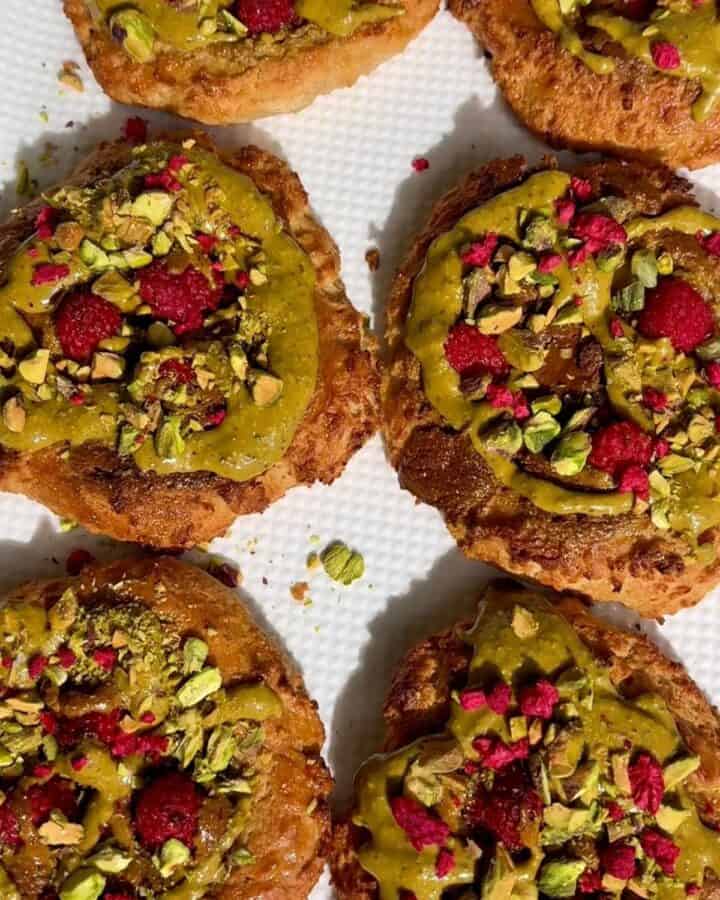 plate of pistachio raspberry breakfast buns in the sunlight, all topped with chopped pistachios, pistachio butter and raspberries