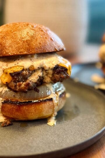 thick, juicy burger sitting on a plate, atop a crispy caramelized onion, sandwiched between two toasted buns and dripping in punchy burger sauce