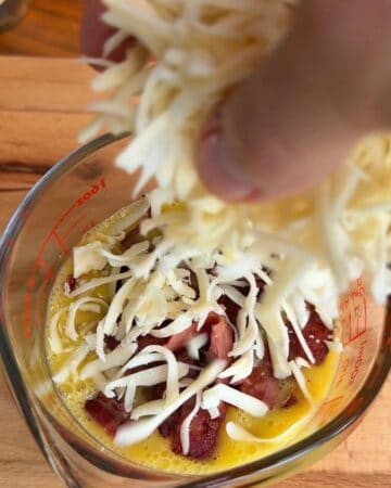 whisked egg, bacon and cheese being added to a bowl in prep for the making of a bacon, egg, and cheese tortilla muffin