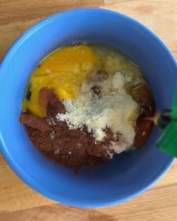 egg, almond flour, cocoa, maple syrup being added to a mixing bowl in prep for a high-protein microwave chocolate lava cake