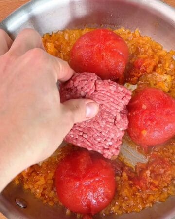 ground beef being added to a pan of blistering tomatoes, sautéed onions, for the making of one-pan cheesy tomato beef rigatoni with shredded parmesan and parsley