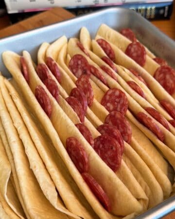 tortillas folded in a baking dish with pepperoni layered on top, in prep for crispy, cheesy, tortilla pull apart pizza bread