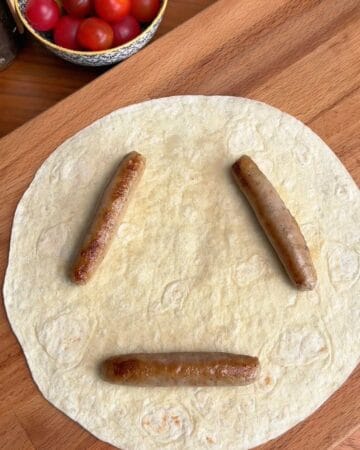 a tortilla with three breakfast sausages on it, ready to be rolled into the crust for a breakfast pizza with sausage stuffed tortilla crust