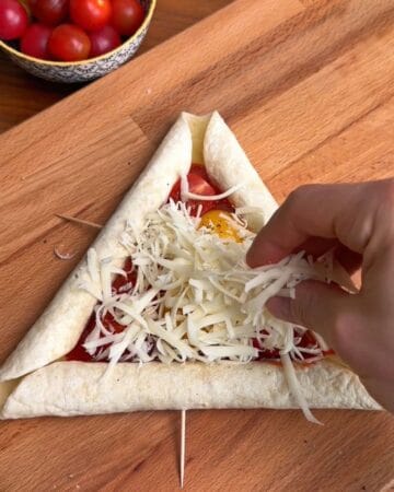 shredded cheese being spread on top of cherry tomatoes, egg yolks and marinara in prep for the making of a breakfast pizza with sausage stuffed crust