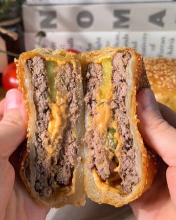 juicy cheeseburger wellington sliced in half showing layers of burger patty cheddar, pickles, caramelized onions and puff pastry