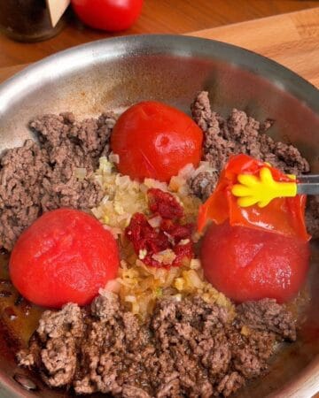 a pair of tongs removing the skins on tomatoes in a pan of beef and seasonings for pasta sauce