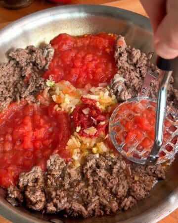 tomatoes being crushed in a pan with ground beef and seasonings for a creamy ground beef pasta made with fresh tomatoes