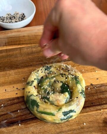 rice paper stuffed with spanakopita filling rolled into a tight spiral, being sprinkled with sesame seeds