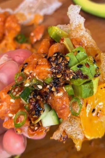 a bite of a spicy salmon tostada, made with rice paper, spicy salmon, green onion, cucumber and sesame seeds