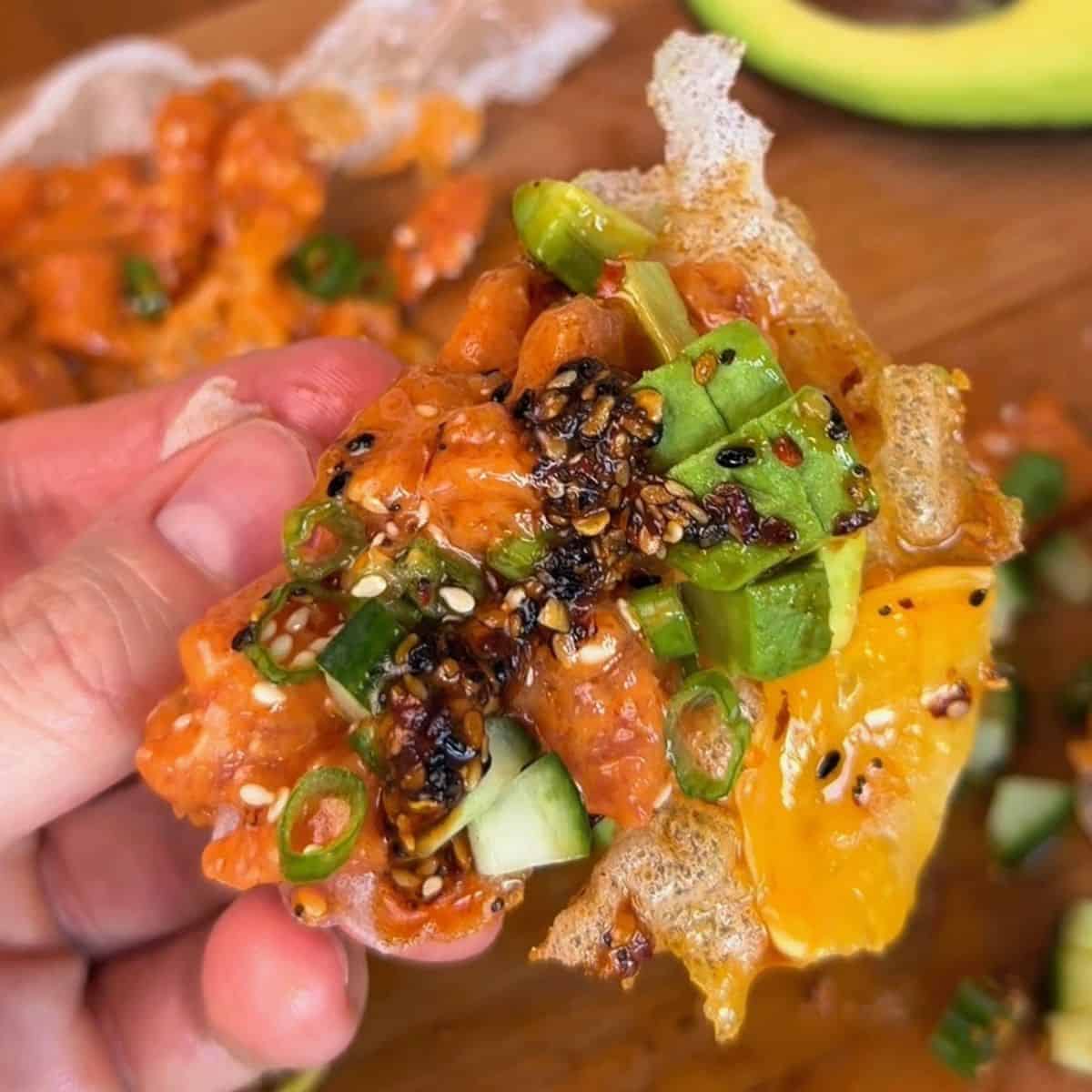 a bite of a spicy salmon tostada, made with rice paper, spicy salmon, green onion, cucumber and sesame seeds