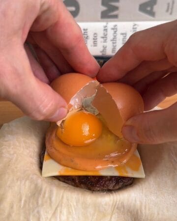 egg being layed on top of cheese, burger sauce, and a sausage patty, layered in a puff pastry, for a breakfast sandwich wellington