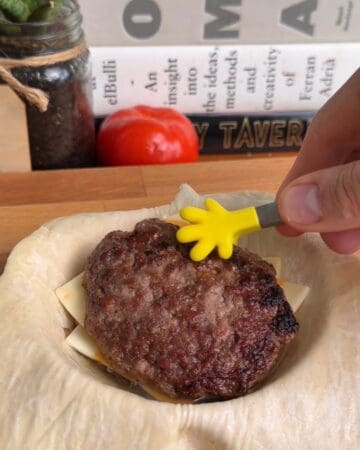 a sausage patty being laid on top of a stack of cheese, egg, burger sauce, and sausage, for the making of a breakfast sandwich wellington
