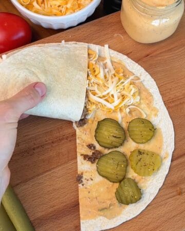a tortilla, layered with burger sauce, ground beef, cheese and pickles, being folded using the triangle method for the making of a crispy smashburger quesadilla