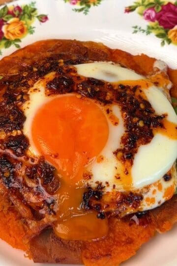 a crispy smashed sweet potato on a plate, layered with avocado, a funny egg, and topped with chili crisp