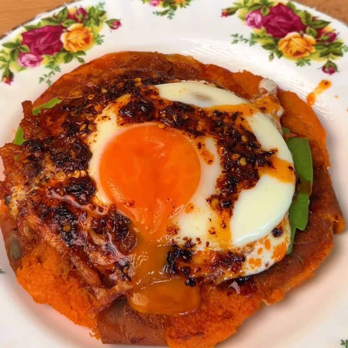 a crispy smashed sweet potato on a plate, layered with avocado, a funny egg, and topped with chili crisp