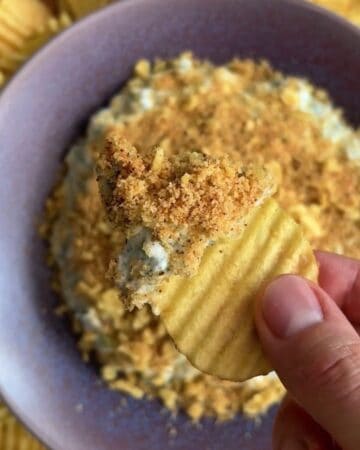 Hand holding a potato chip fried pickle dip in a shallow dish with crispy panko and chip topping surrounded by kettle chips for scooping