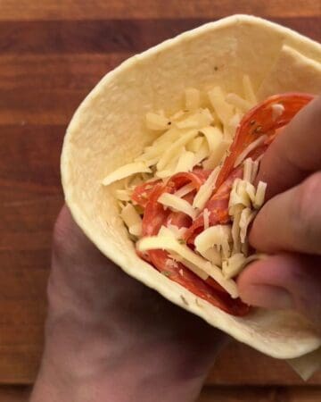 folded tortilla being filled with pepperoni, shredded cheese and italian seasoning for a pepperoni pizza pocket