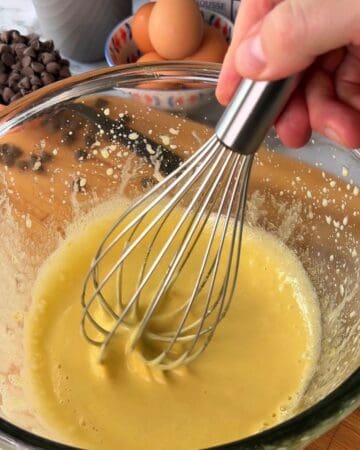 Whisk blending two-bite brownie batter in a glass bowl with eggs and chocolate chips nearby.
