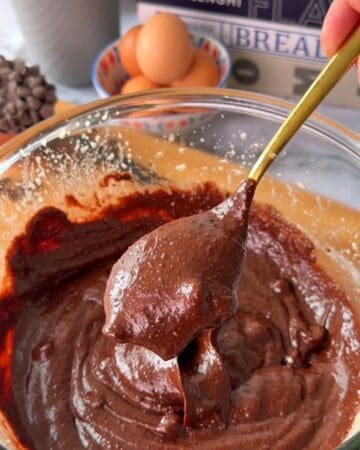 Chocolate two-bite brownie batter in a glass bowl with a spoon, eggs, and baking ingredients in the background.