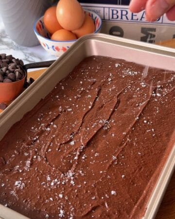 Two-bite protein brownie batter in a baking pan with baking ingredients in the background.