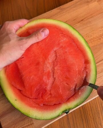 Person scooping watermelon flesh from a watermelon half on a wooden surface.