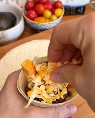 Close-up of hand adding shredded cheese to taco cone with ground beef.