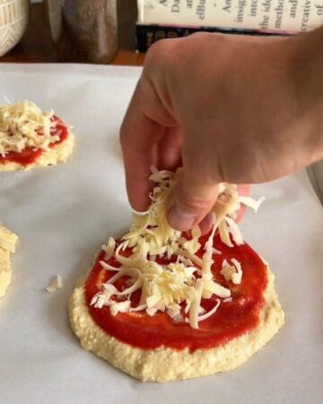 Hand adding shredded cheese on pizza dough with tomato sauce.