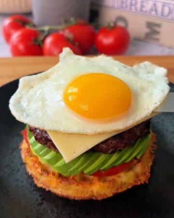 Delicious breakfast burger with fried egg, avocado, tomato, and cheese on a potato waffle bun.