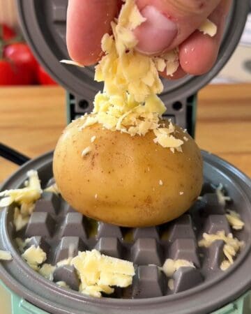 Baked potato with shredded cheese being added on top, on a mini waffle maker