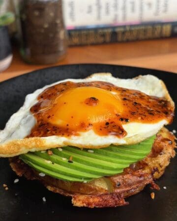 Delicious breakfast stack with fried egg, avocado slices, and crispy potato waffle on a black plate.