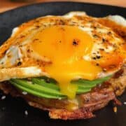 Close-up of a breakfast stack with fried egg, avocado slices, and crispy potato waffle on a black plate.