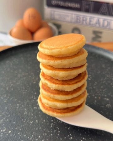 Stack of golden-brown pancakes on a black plate with eggs in the background.