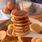 Stack of fluffy pancakes drizzled with syrup, served with eggs in the background.