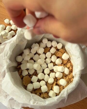 Person adding marshmallows to s'mores cheesecake base with graham crackers.