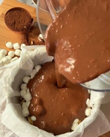 Pouring rich chocolate s'mores cheesecake batter into pan with marshmallow toppings